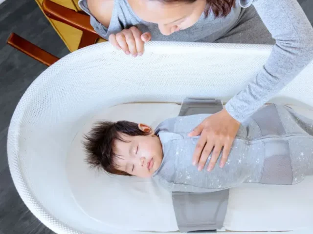 An adult leans over a baby sleeping in a white bassinet gently resting a hand on the baby’s chest. The baby is swaddled in a gray wrap.
